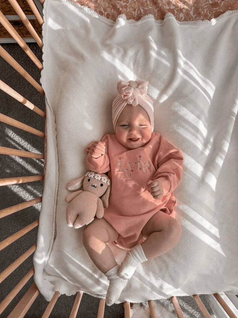 Adorable baby with personalised baby keepsakes, smiling in a cozy crib wearing a cute pink outfit and headband.