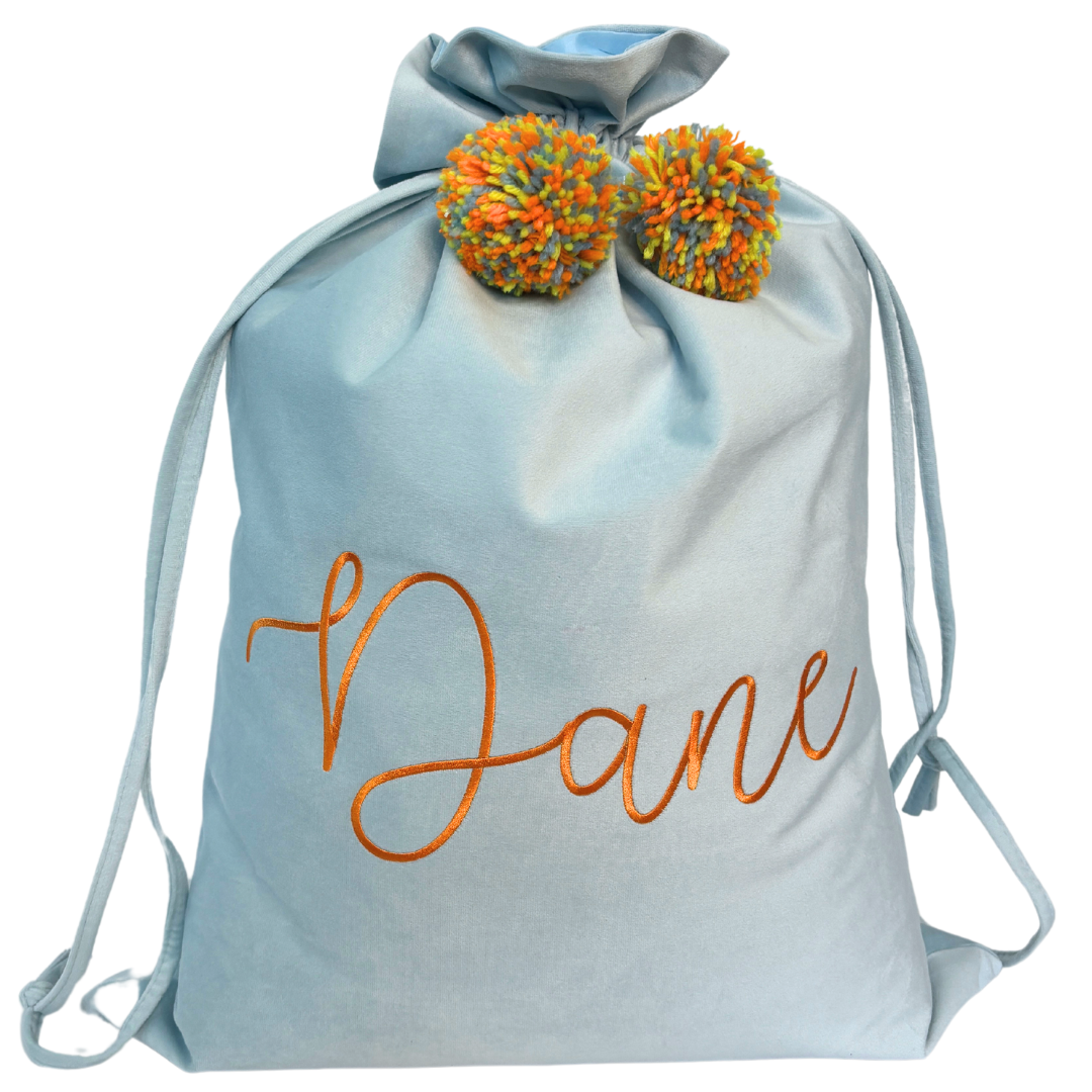 Personalised light blue velvet Santa sack with orange embroidery reading ‘Dane’ and colourful orange and yellow pom poms.
