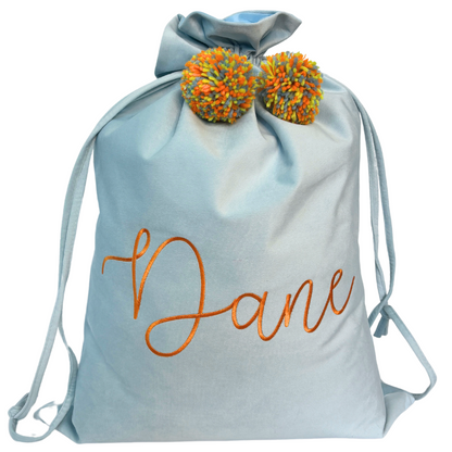 Personalised light blue velvet Santa sack with orange embroidery reading ‘Dane’ and colourful orange and yellow pom poms.