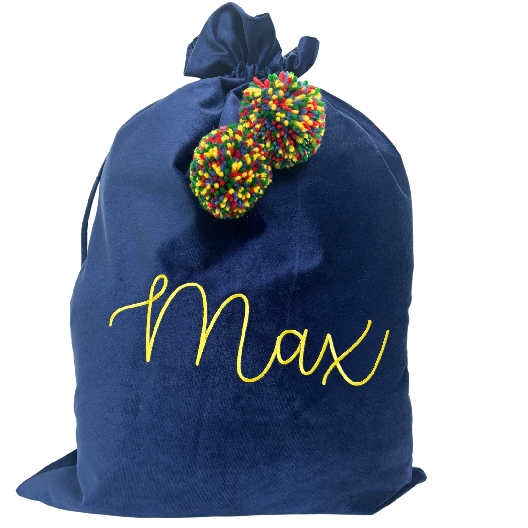 Personalised navy velvet Santa sack with bright yellow embroidered name ‘Max’ and rainbow pom poms in red, green, yellow, and blue