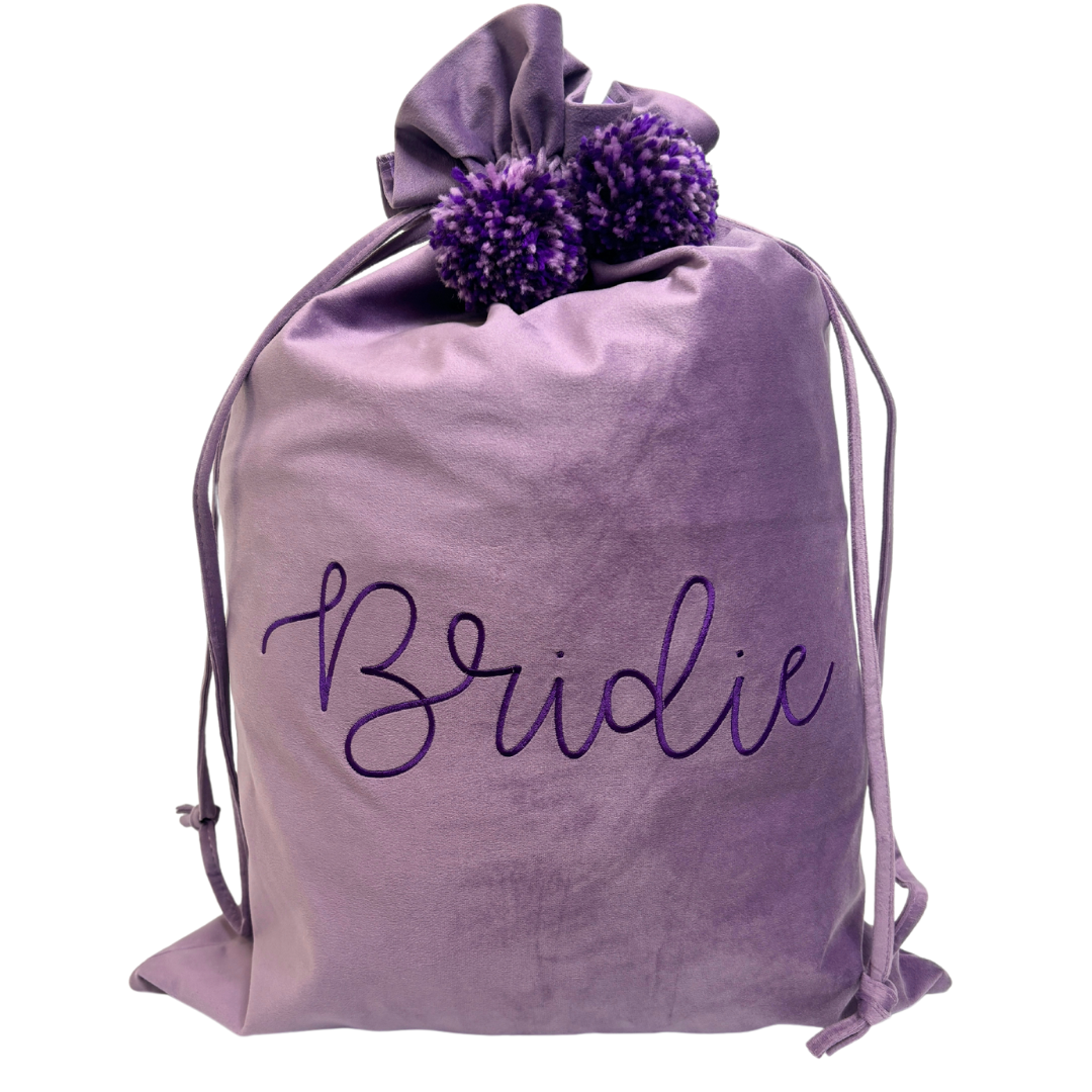 Personalised lavender velvet Santa sack with deep purple embroidery reading ‘Bridie’ and matching purple pom poms.