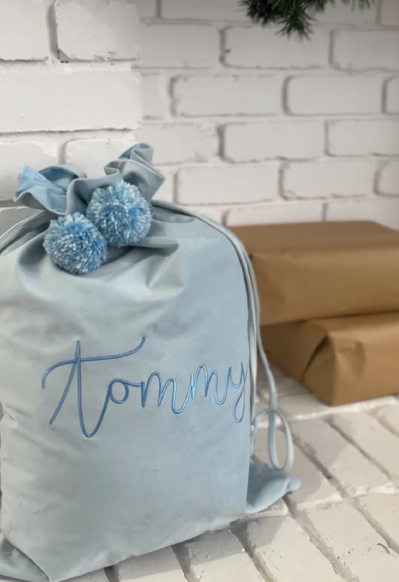 Light blue velvet Santa sack embroidered with name Tommy in blue thread, finished with fluffy blue pom poms beside Christmas presents