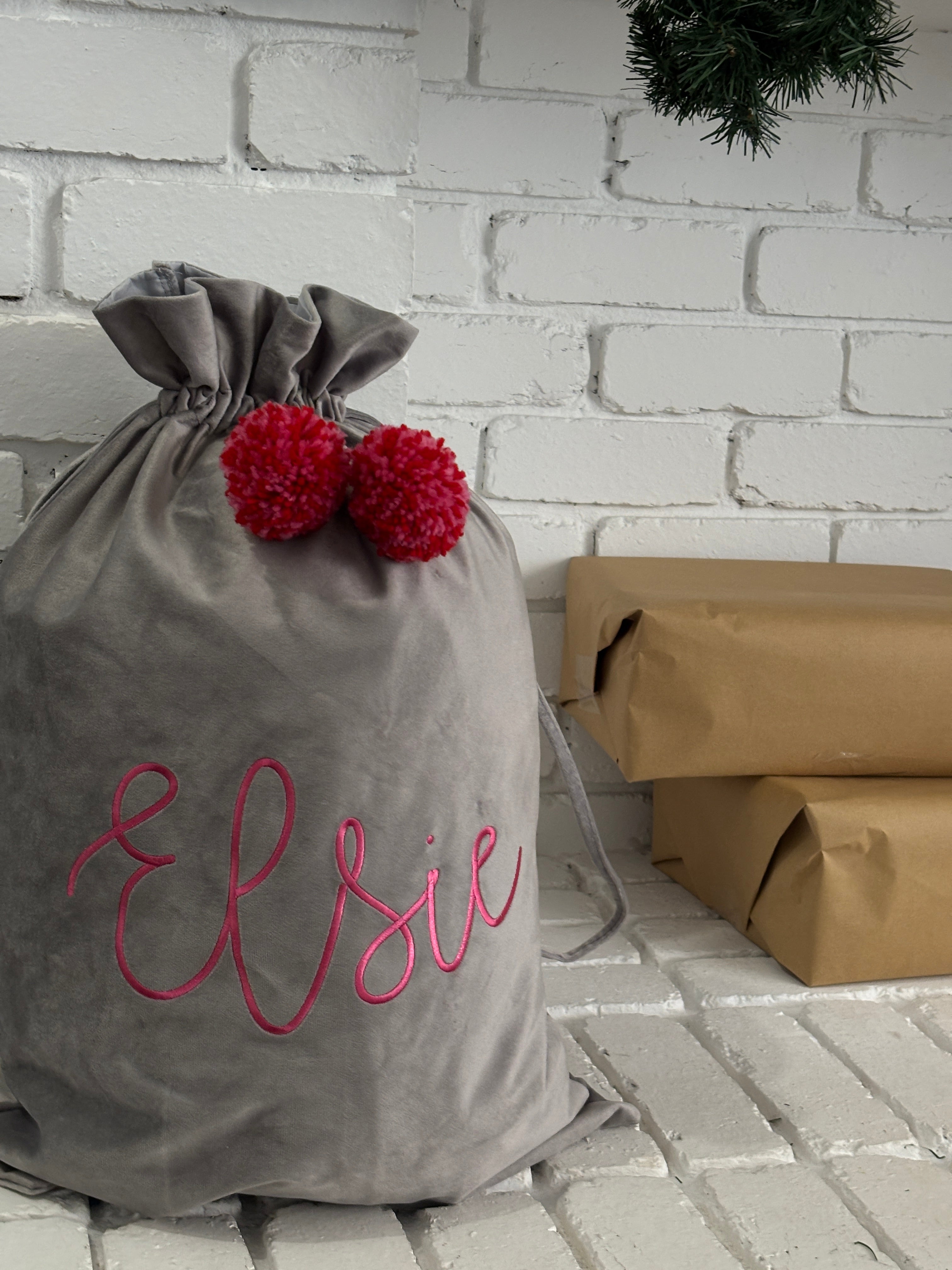 Grey velvet Santa sack embroidered with name Elsie in pink thread and topped with red pom poms, sitting by Christmas gifts under greenery.