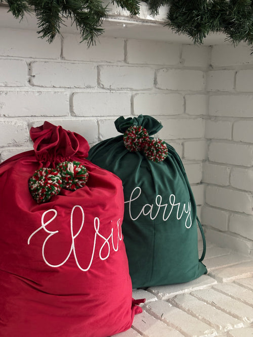 Red and green personalised velvet Santa sacks embroidered with names Elsie and Harry, styled under a white brick fireplace with festive greenery.