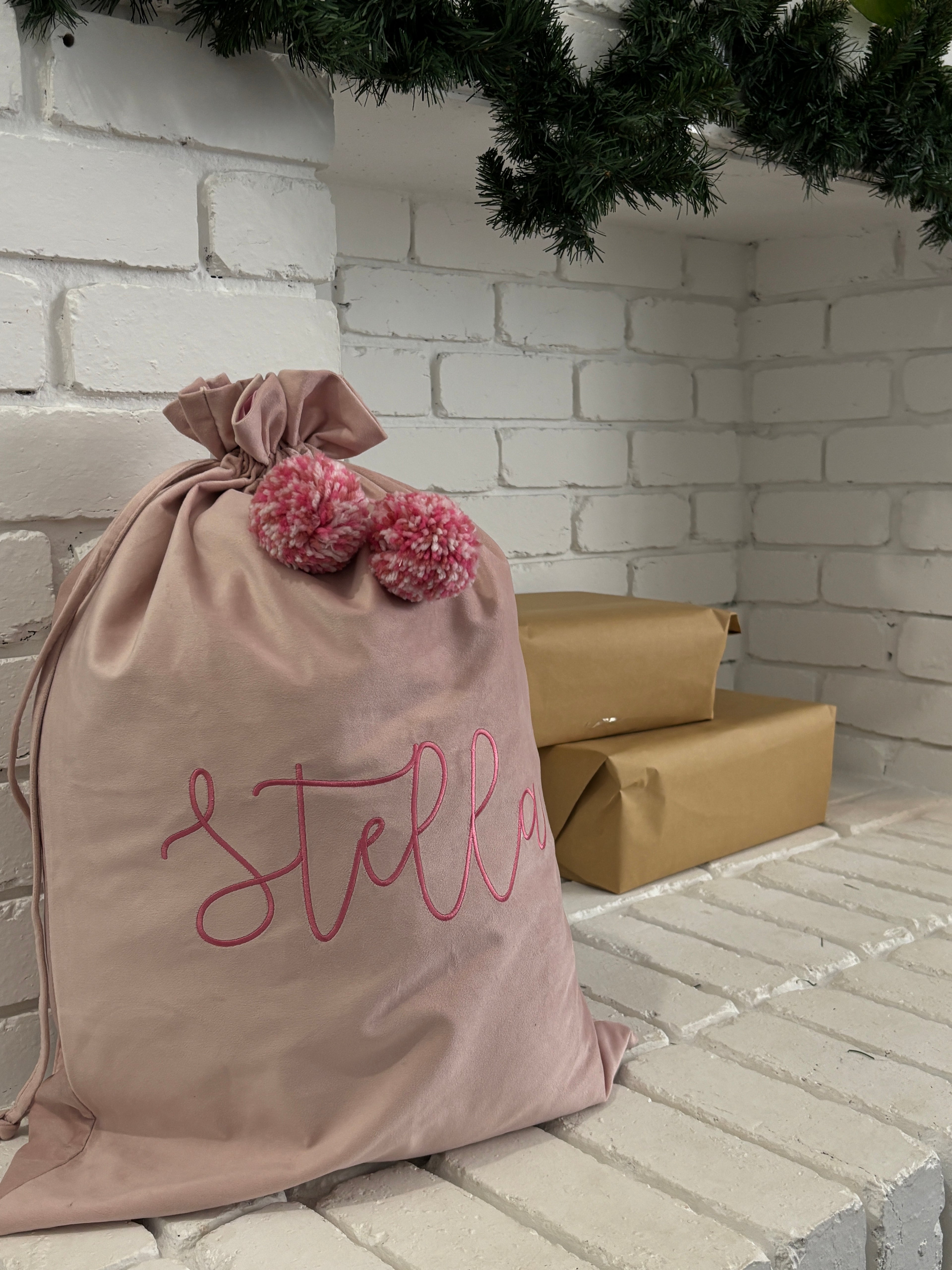 pink velvet Santa sack with embroidered name Stella and soft pink pom poms, styled on a white brick fireplace beside brown wrapped presents.