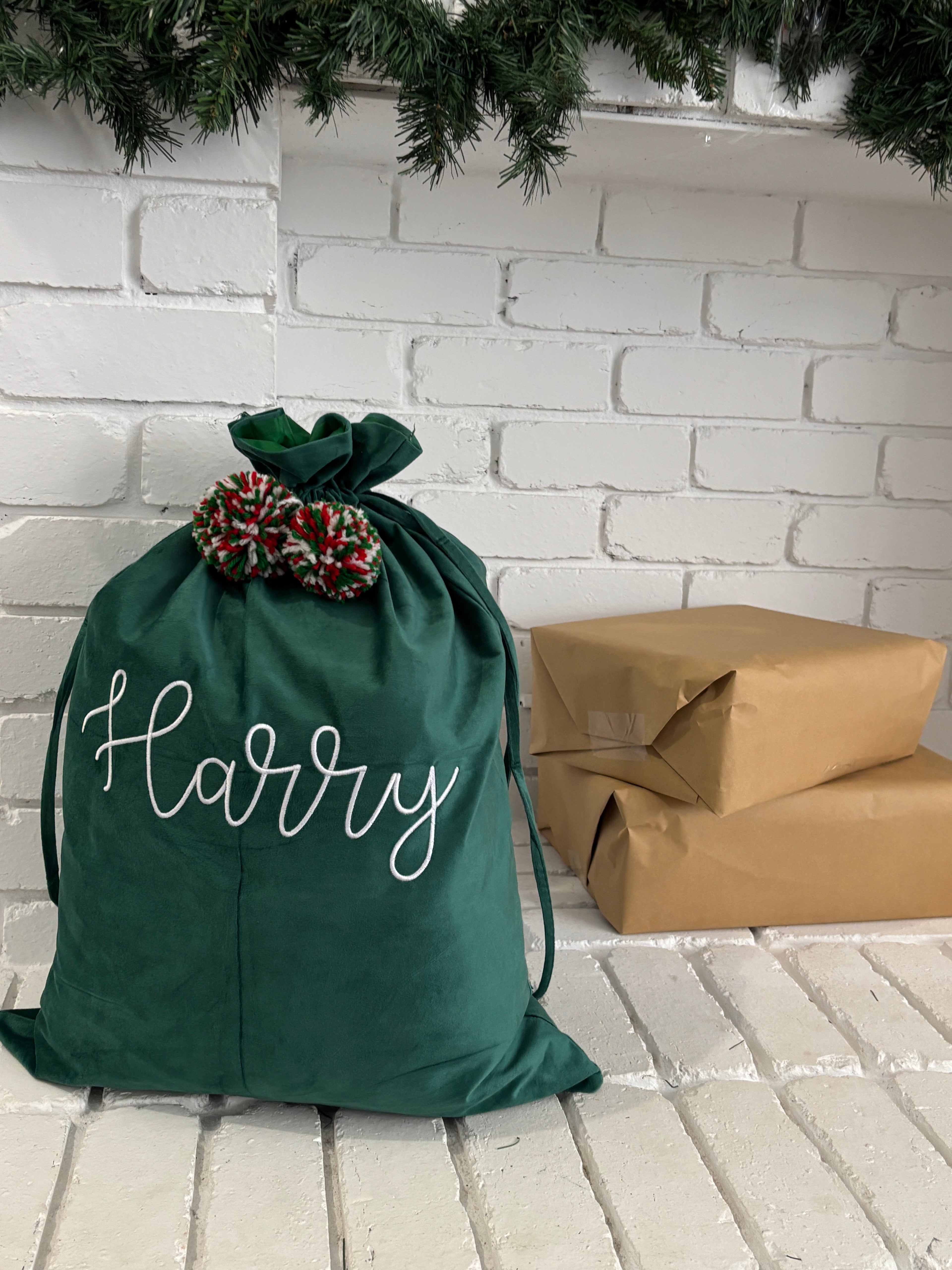 Green velvet Santa sack embroidered with name Harry, displayed beside brown paper–wrapped Christmas gifts and festive garland.