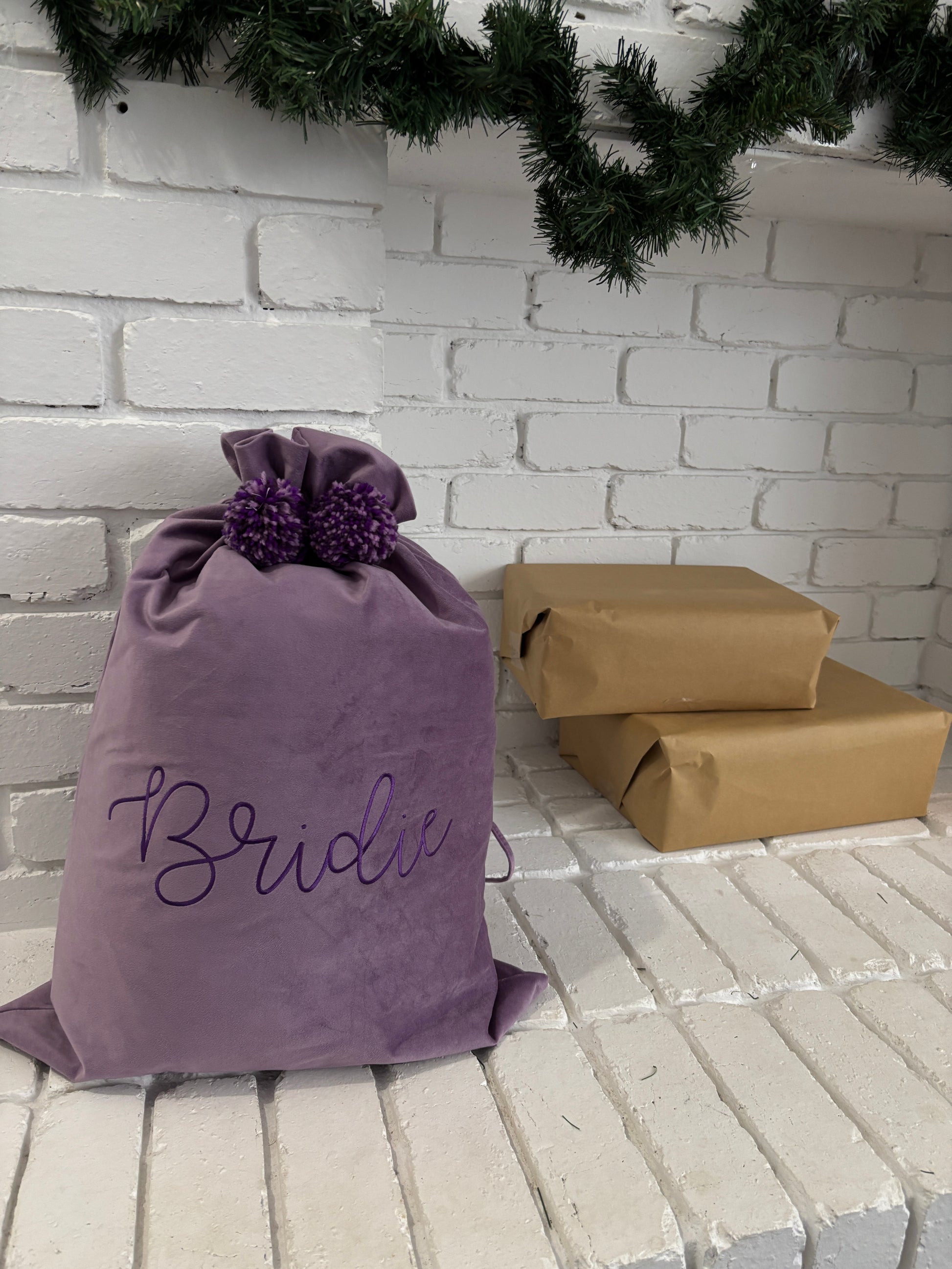 Purple velvet Santa sack with embroidered name Bridie and matching pom poms, placed under a Christmas garland beside wrapped presents.