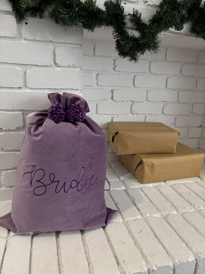 Purple velvet Santa sack with embroidered name Bridie and matching pom poms, placed under a Christmas garland beside wrapped presents.